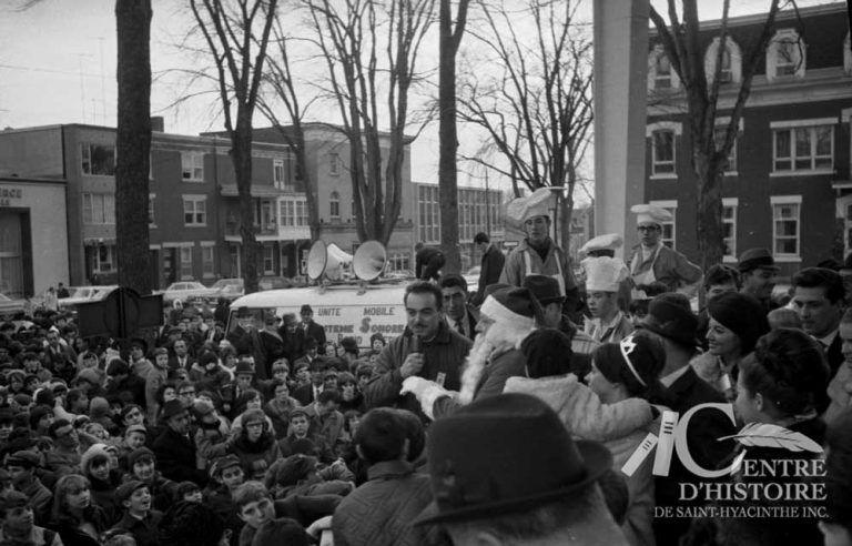 HO! HO! HO!1966. - Fonds Raymond Bélanger, CH548.En 1966, le défilé se termine au parc Dessaulles. L'animateur Henri Bergeron et le Père Noël adressent quelques mots à la foule. Cette année-là, les journaux maskoutains indiquent qu'environ 30 000 personnes assistent à l'événement.